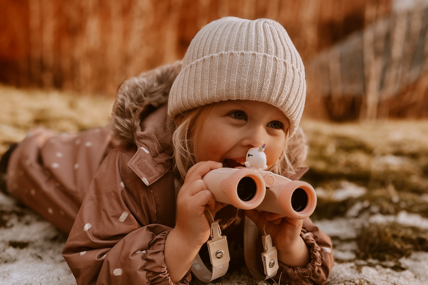 The girl is lying on the ground, smiling and looking through her binoculars.

